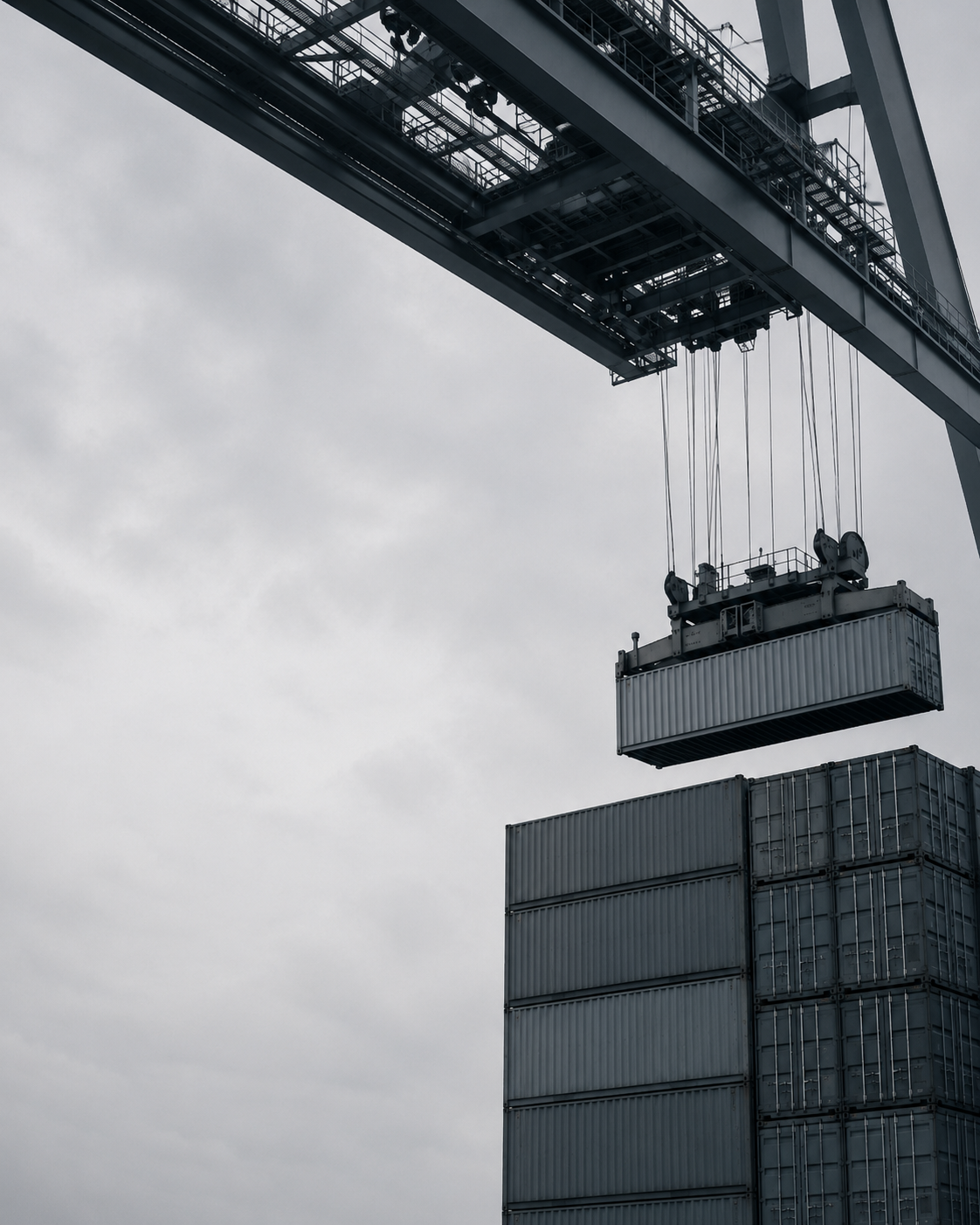 A port gantry crane lifts a shipping container above a stack of container units at a freight yard.