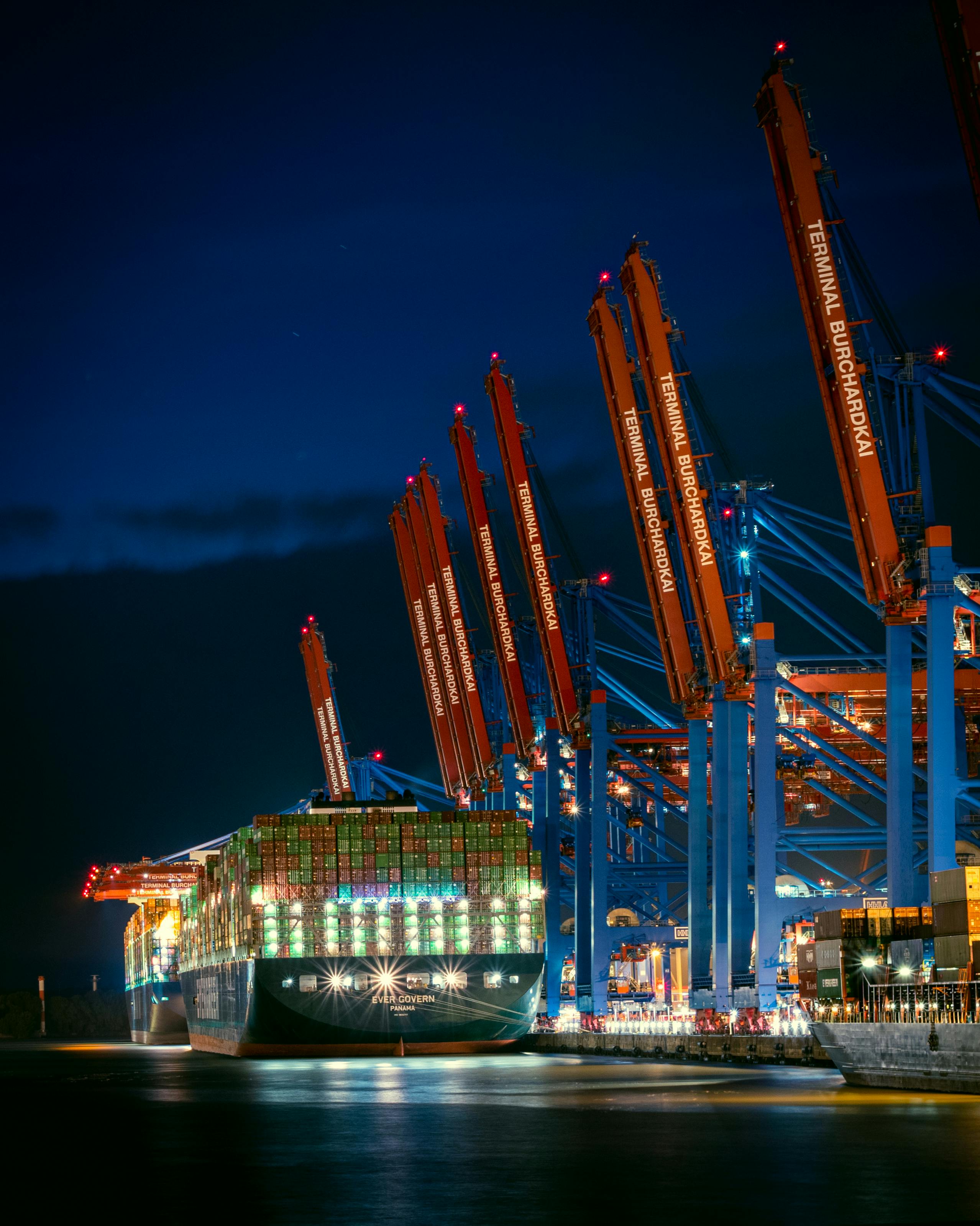 A large Evergreen container vessel docked at a busy port terminal under illuminated gantry cranes at night.