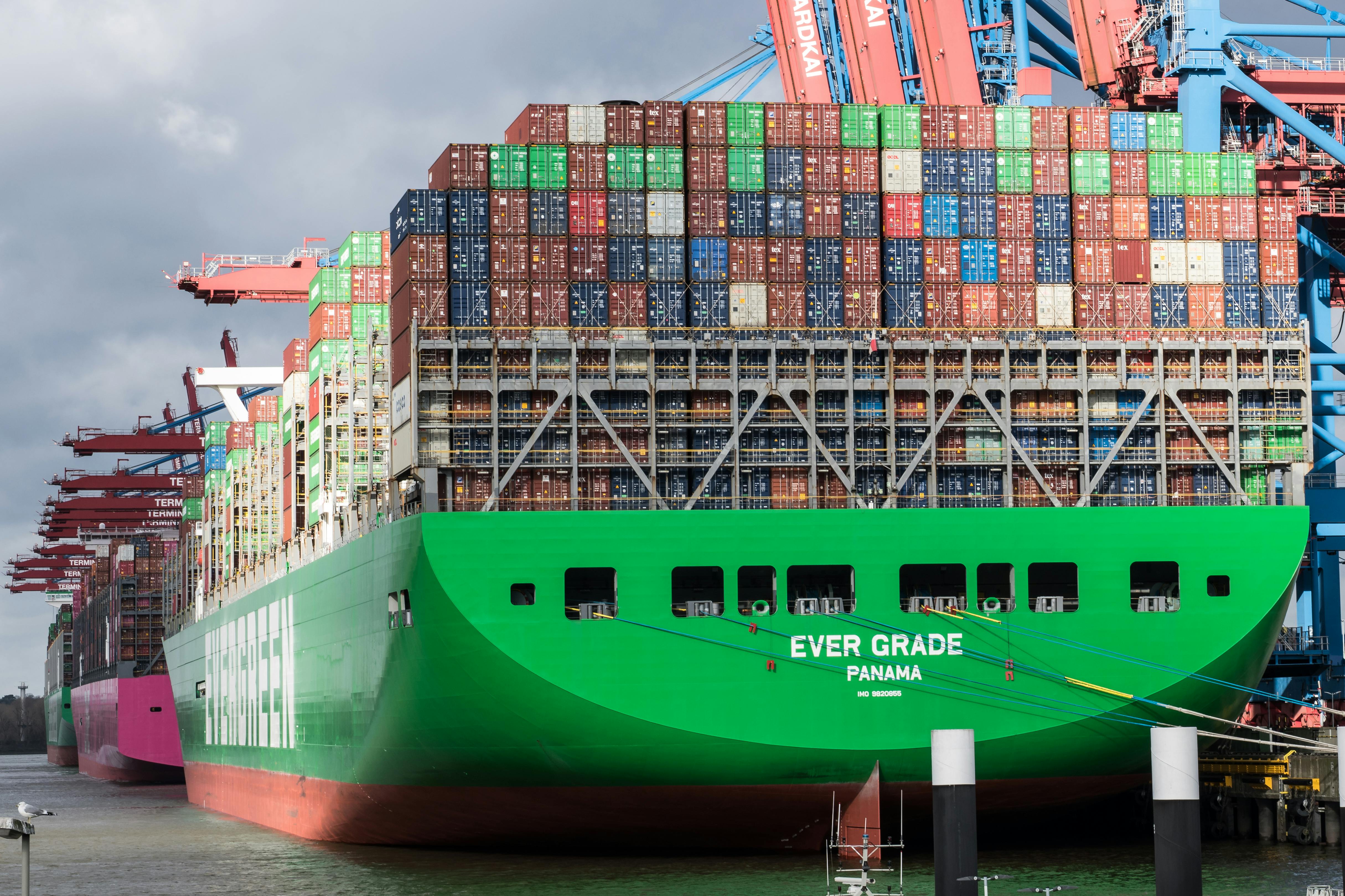 A large Evergreen container ship named EVER GRADE loaded with colorful shipping containers docked at a port terminal under gantry cranes.