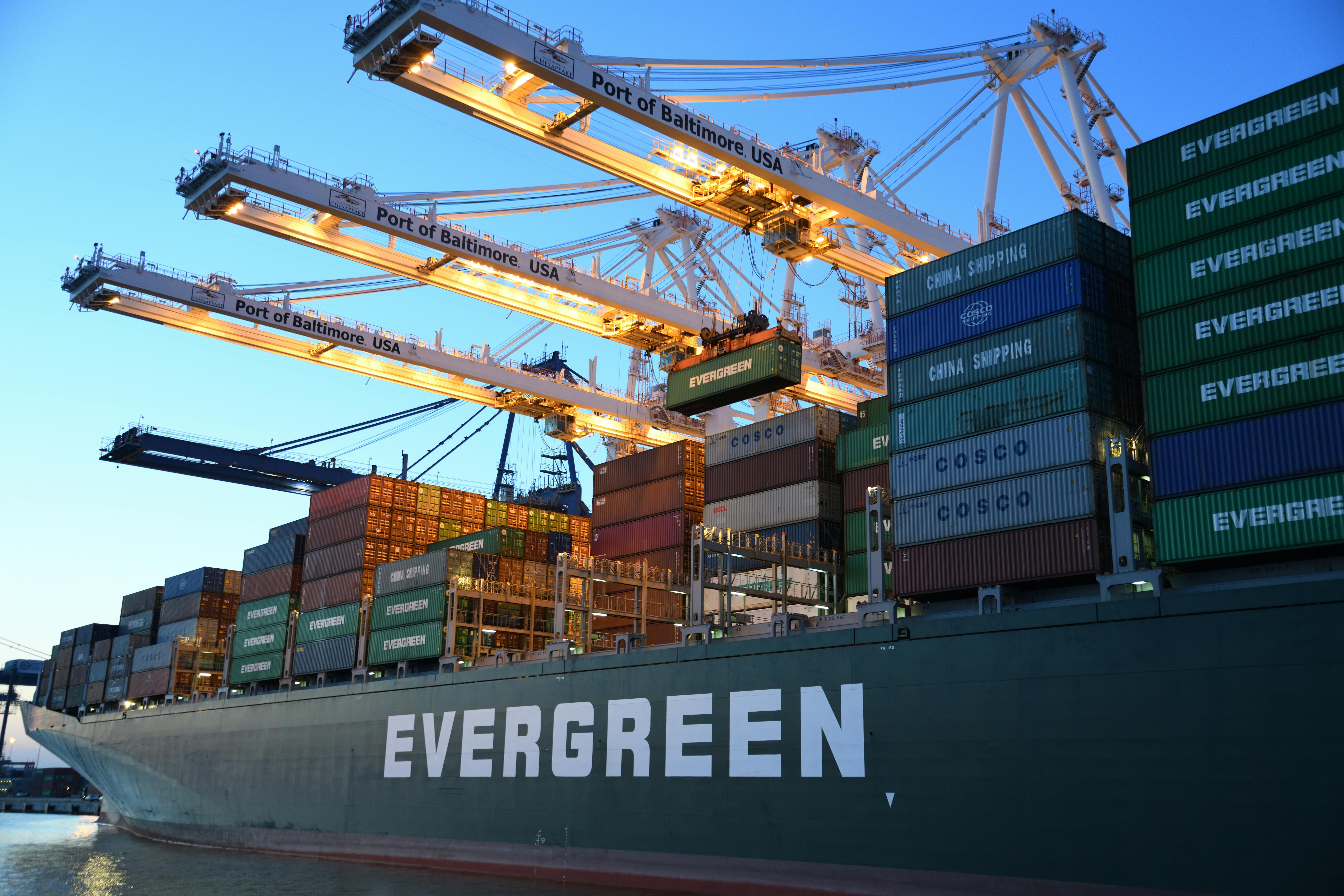 An Evergreen container vessel being loaded and unloaded by large gantry cranes at a port terminal at dusk.