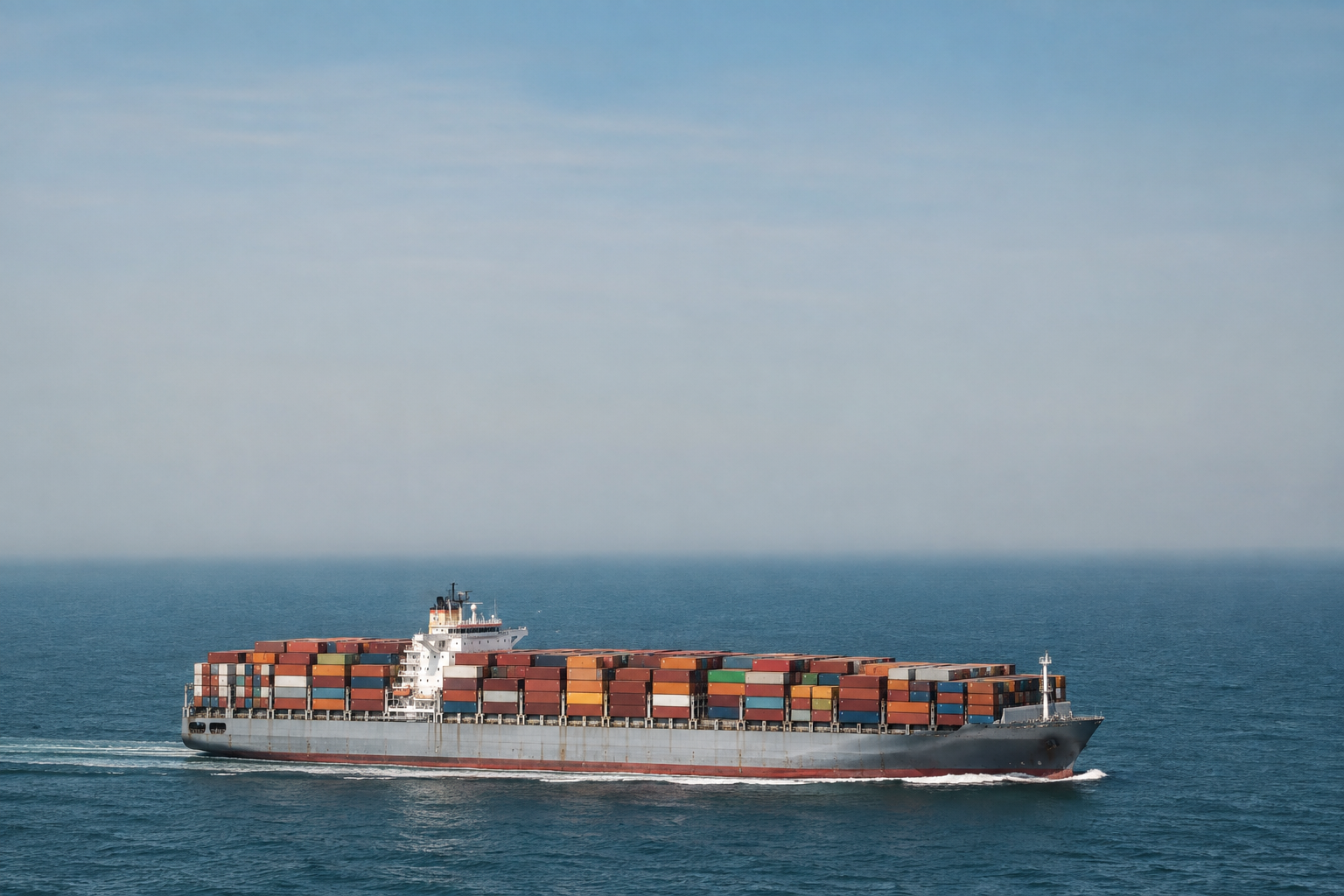 A large container ship heavily loaded with intermodal freight containers navigating across open ocean waters.