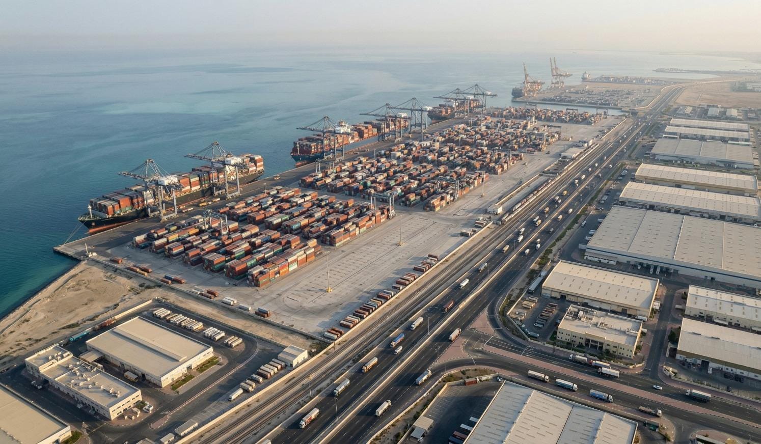 An aerial view of a massive sea port terminal featuring docked container ships, quay cranes, extensive container yards, and adjacent warehouses along a highway.