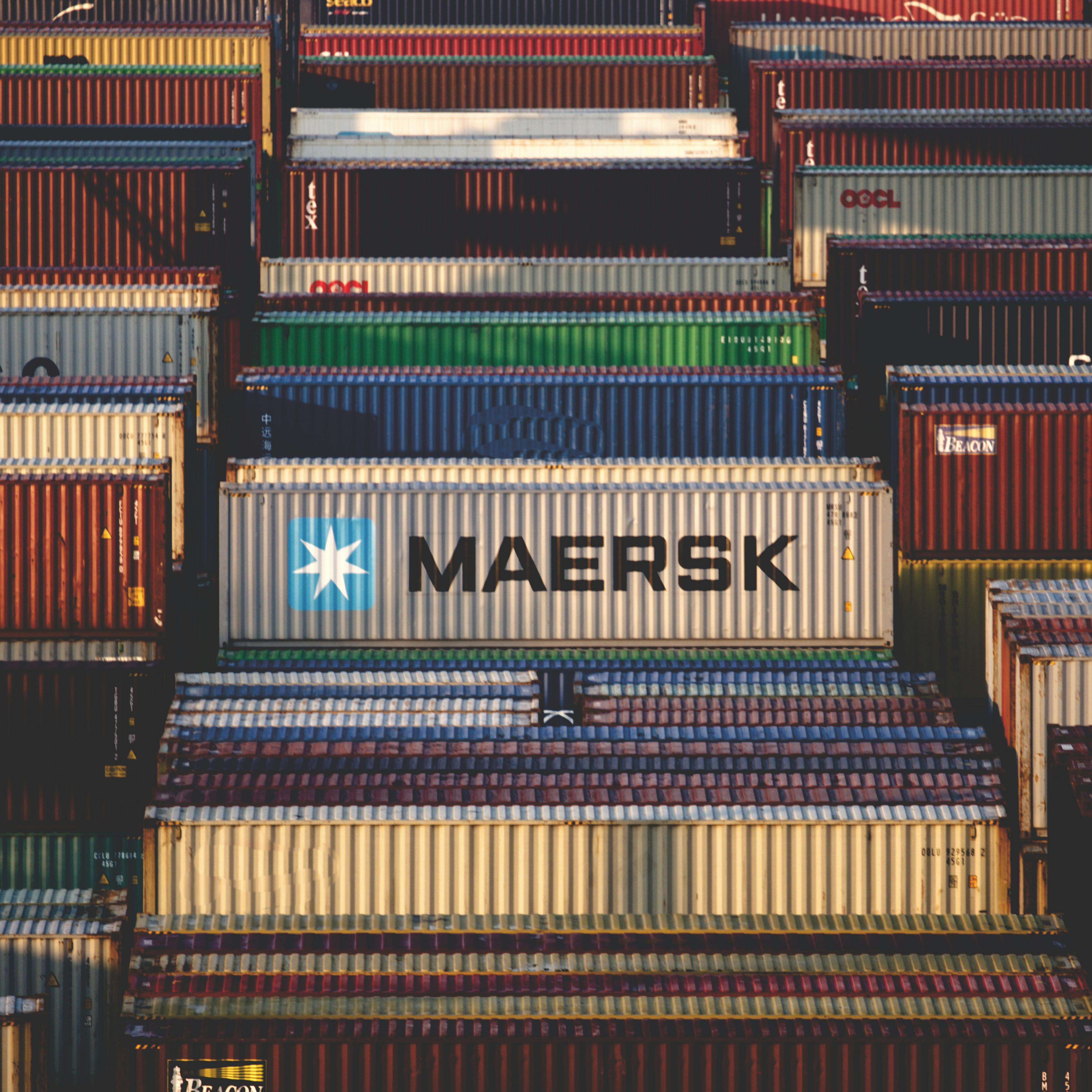 A dense arrangement of stacked intermodal shipping containers in a freight terminal yard featuring prominent branding such as Maersk.